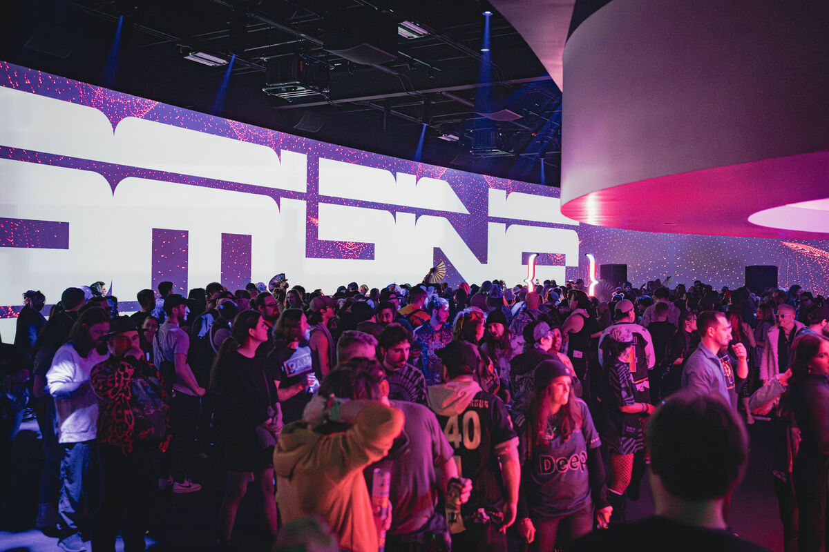 A packed dance floor facing the stage, framed by two lit brace-shaped light sculptures, with a speaker either side of the screen.